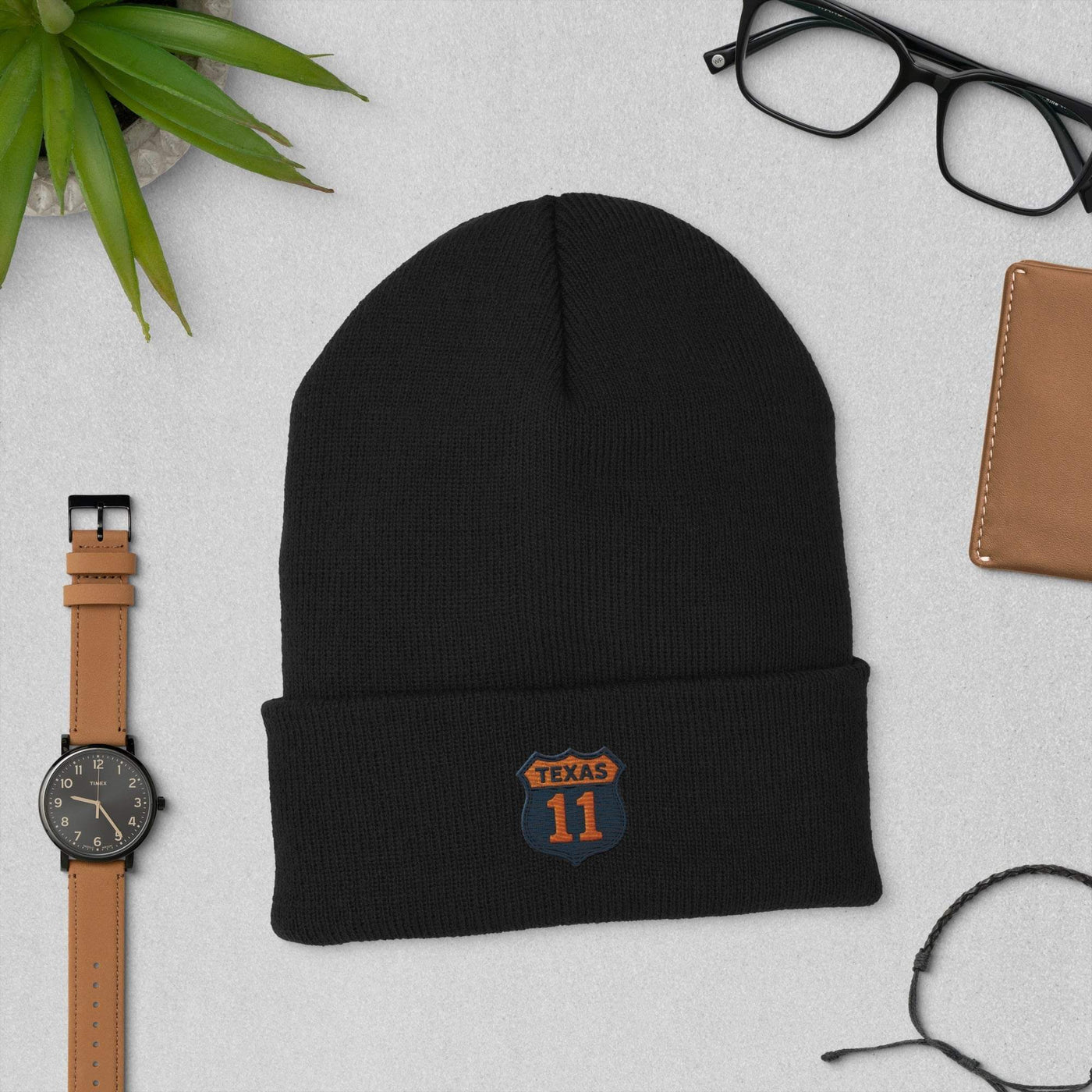 Black cuffed beanie with Texas 11 patch, laid flat with watch, glasses, and wallet on gray surface