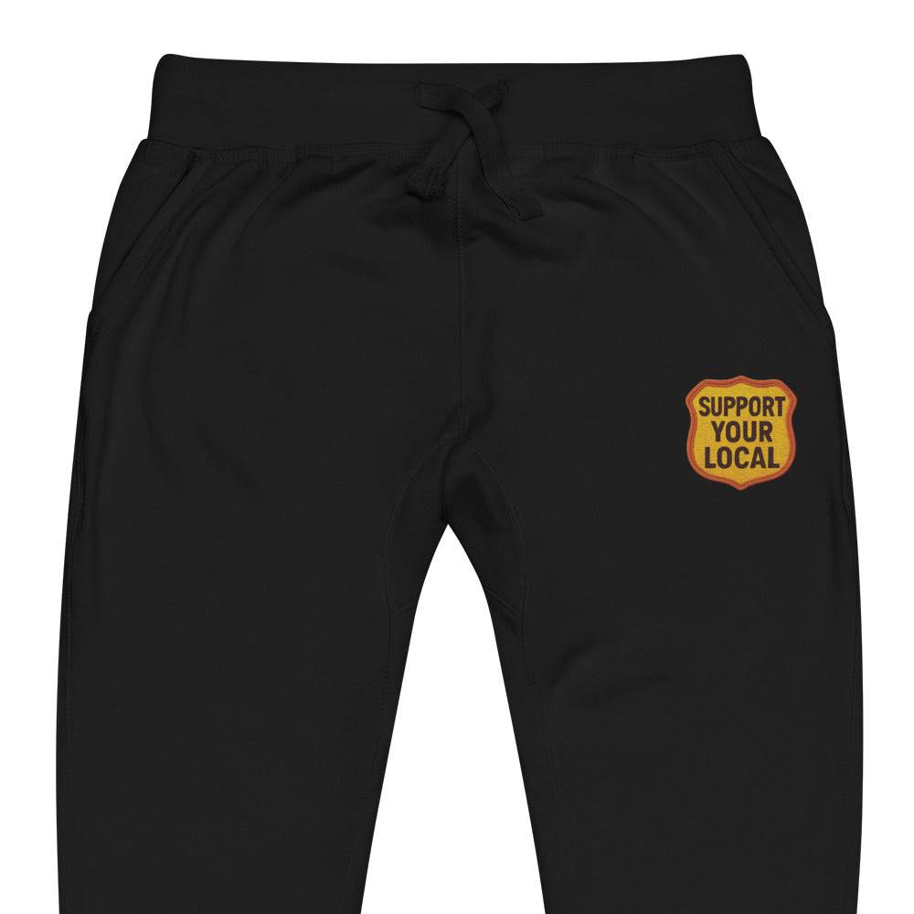 Black unisex fleece sweatpants with elastic waistband, drawstrings, front pockets, and "Support Your Local" patch on thigh.